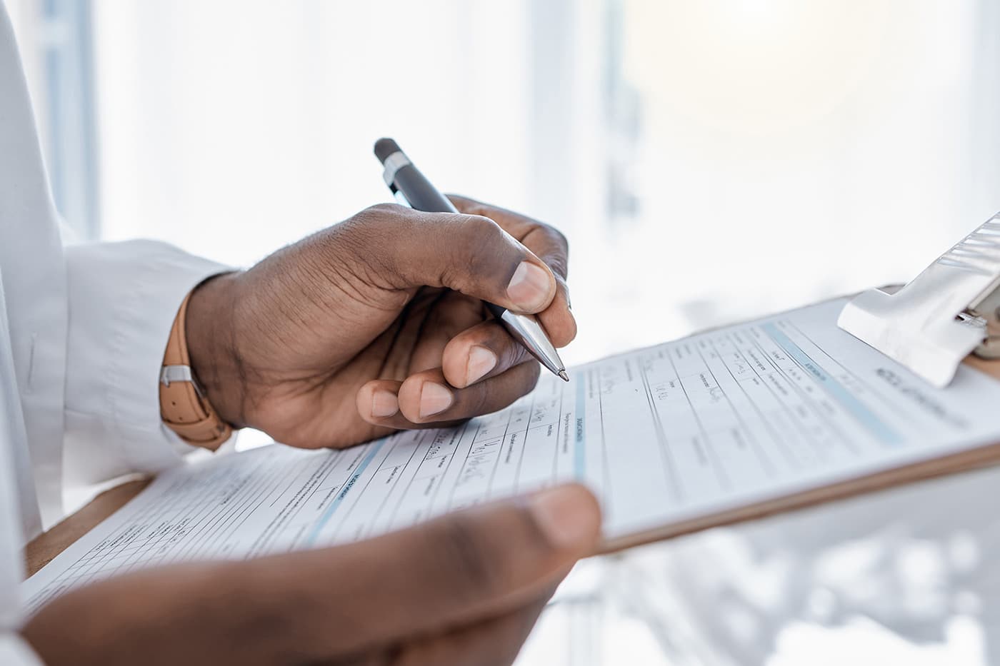 A close-up of a clinician's hand writing on a medical form attached to a clipboard.