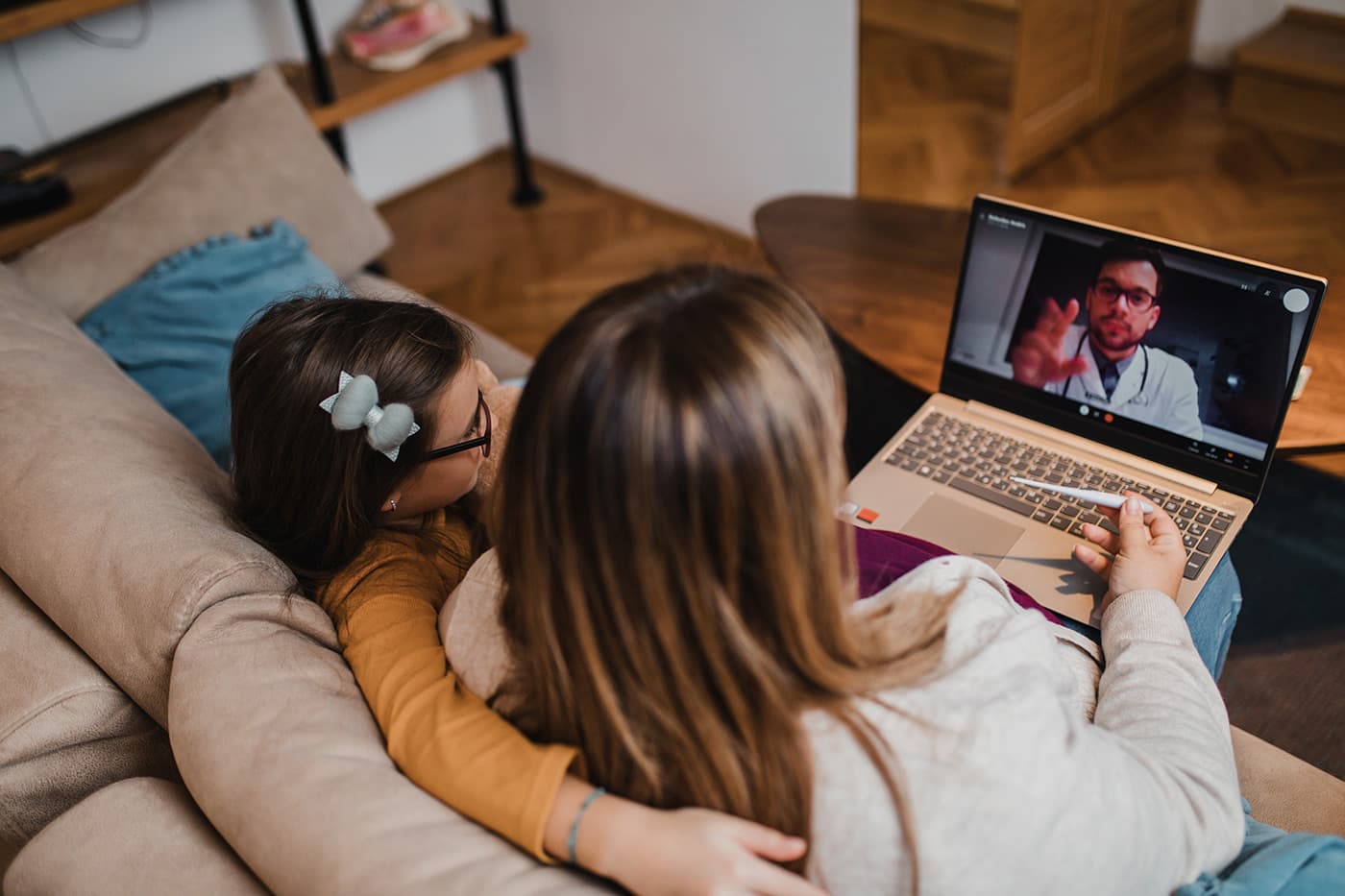 A mother and young child sitting on a couch, viewing a video call with a doctor on a laptop