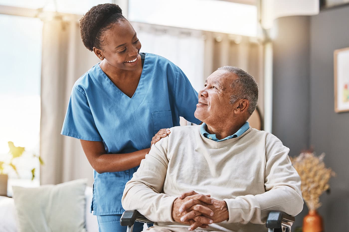 A nurse in blue scrubs smiles warmly at an elderly man seated in a wheelchair in a home care setting.